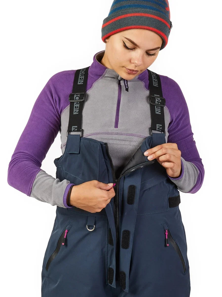 Woman zipping up navy blue ice fishing bib over purple fleece, wearing striped beanie, ready for cold weather